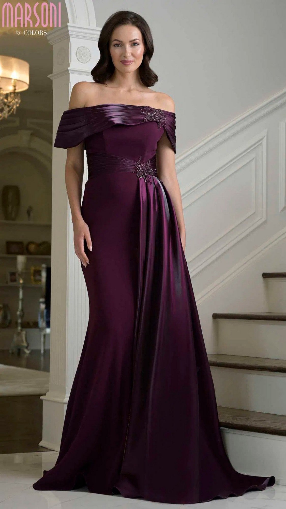 A woman stands on an indoor staircase wearing the Marsoni M347 long formal dress in aubergine, featuring an off-the-shoulder neckline, sheath silhouette, floor-length design, and a decorative accent at the waist.